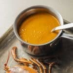 A pot of creamy orange lobster shell sauce next to a lobster head on a baking sheet.