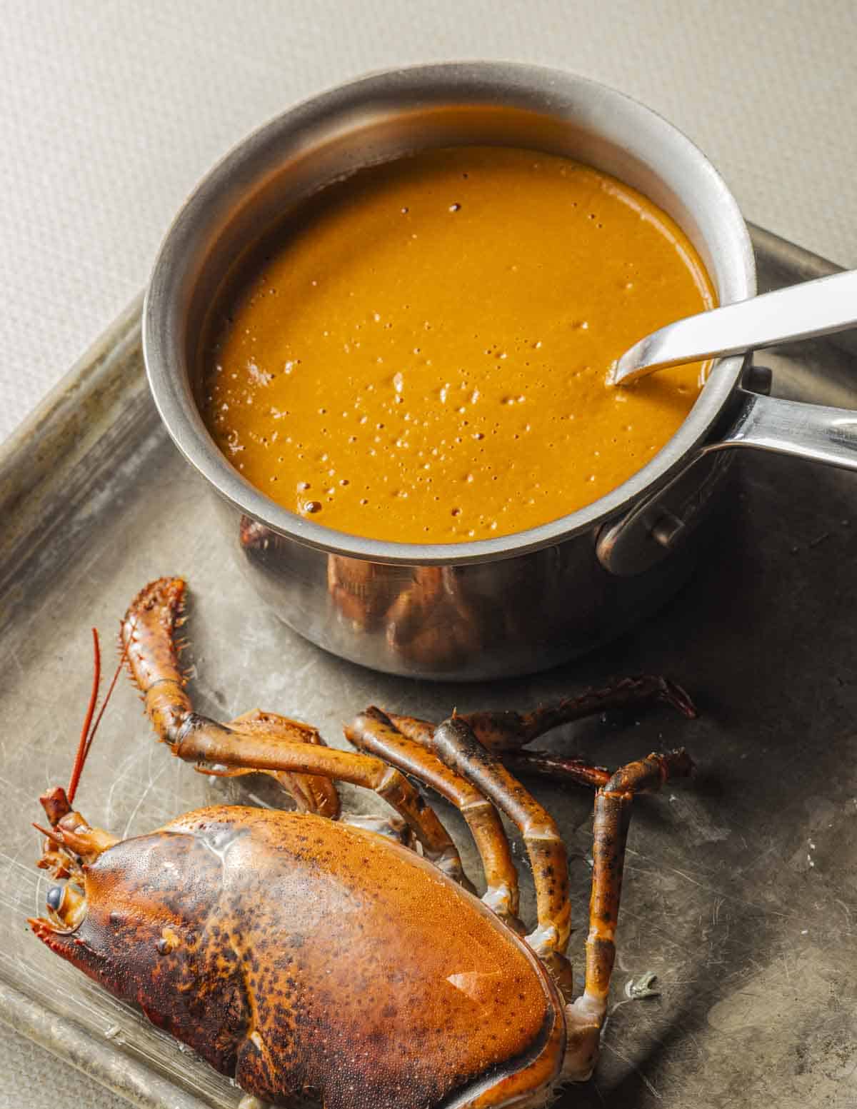 A small pot of orange lobster shell sauce on a baking sheet next to a cooked lobster head.