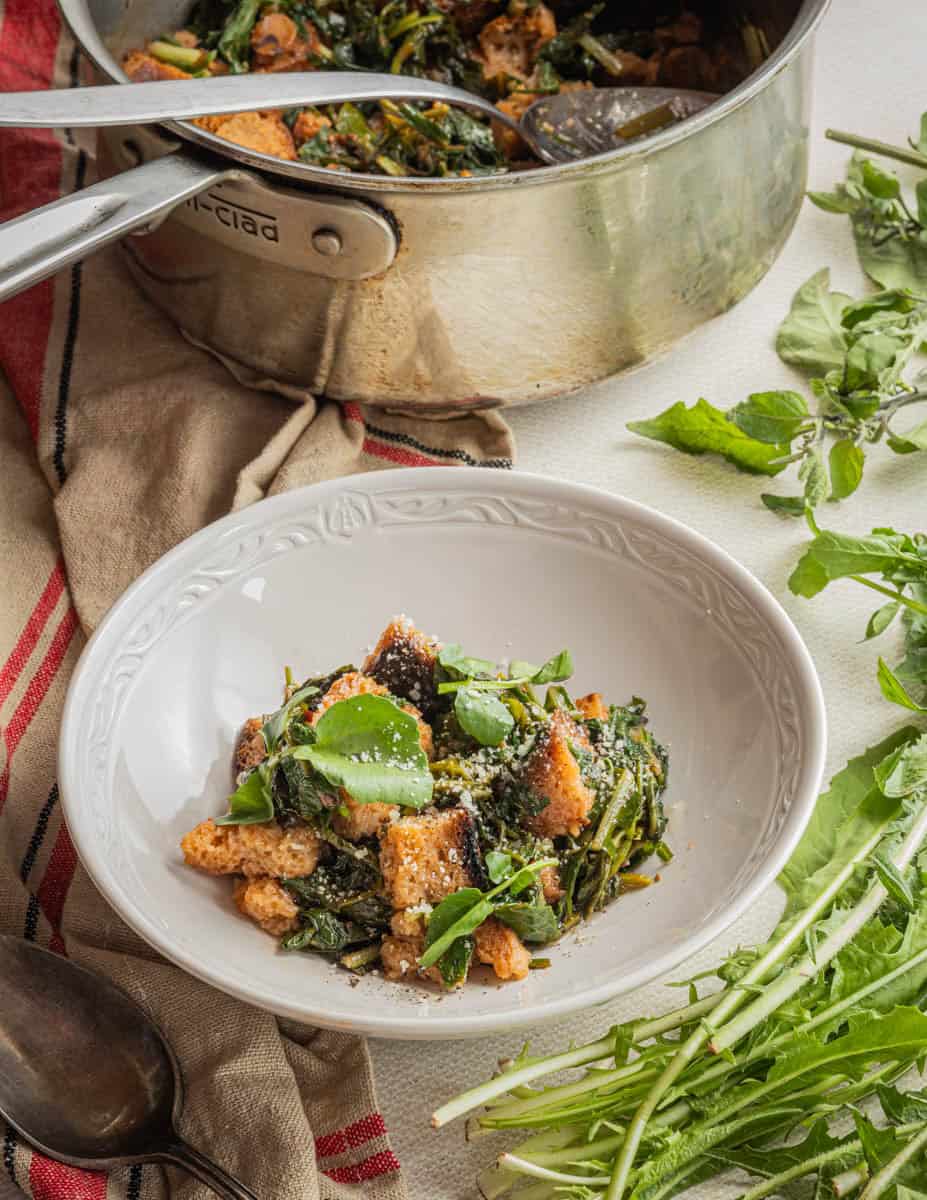 Serving Italian pane cotto in a bowl with a pot in the background surrounded by dandelions, nightshade greens and watercress.