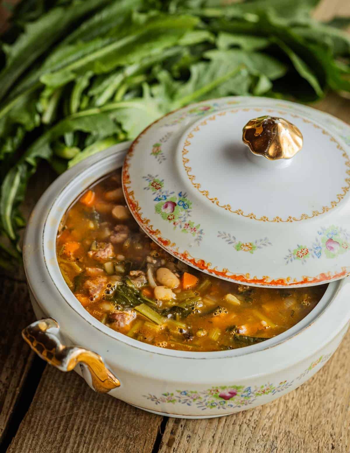 A large terrine of white bean, sausage and dandelion soup in a china soup terrine with a lid with fresh dandelion greens in the background.