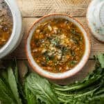 A bowl of sausage, white bean and dandelion soup next to a large soup terrine filled with more soup and surrounded by fresh dandelion greens.