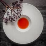 A white bowl showing bright red elderberry vinegar next to a branch of elderberries with fruit on the stems.