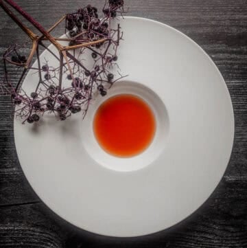 A white bowl showing bright red elderberry vinegar next to a branch of elderberries with fruit on the stems.