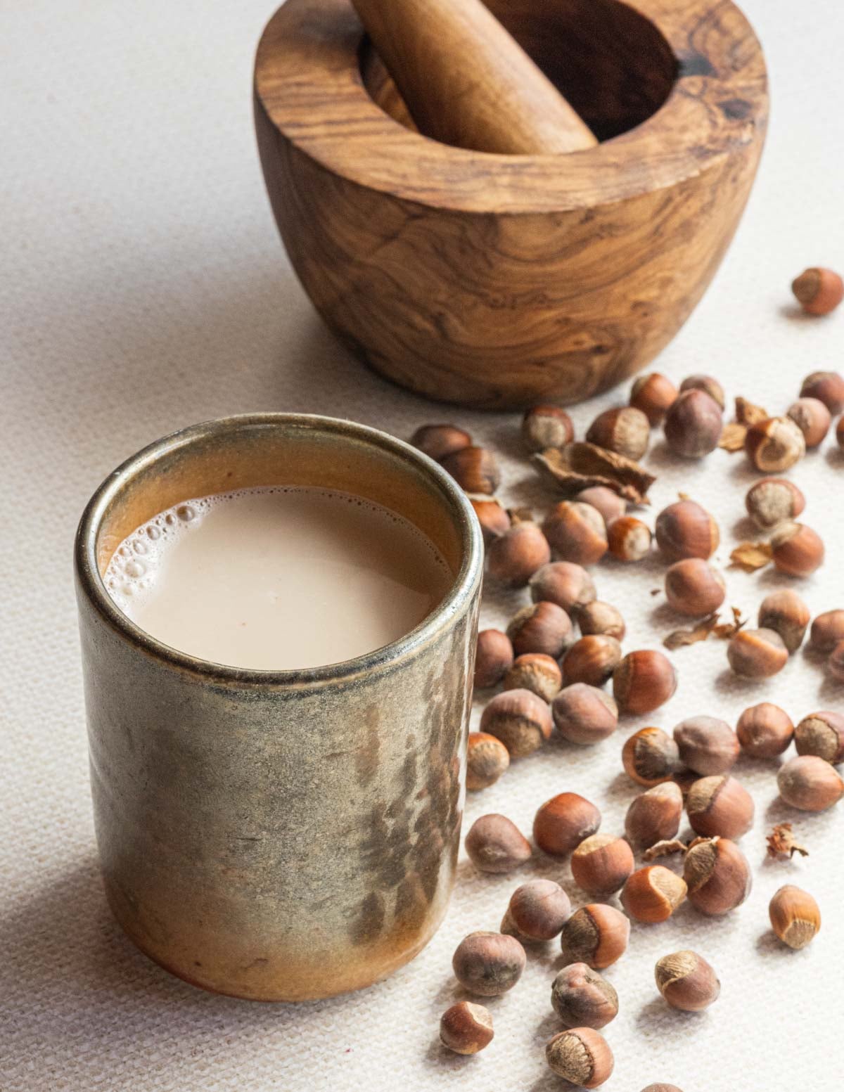 A cup of warm hazelnut milk surrounded by wild hazelnuts in the shell with a mortar and pestle for cracking hazelnuts in the background.