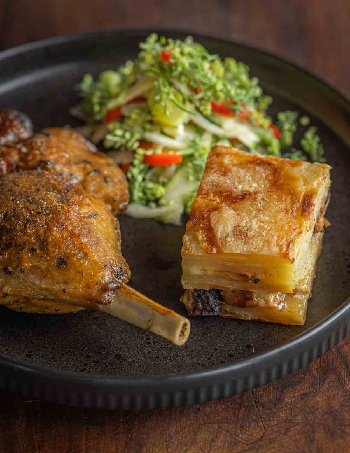 A leg of duck confit on a plate next to a potato gratin and a fennel slaw with virginia pepperweed or peppergrass.
