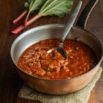 A pan of savory spring rhubarb sauce with rhubarb stems and leaves in the background.
