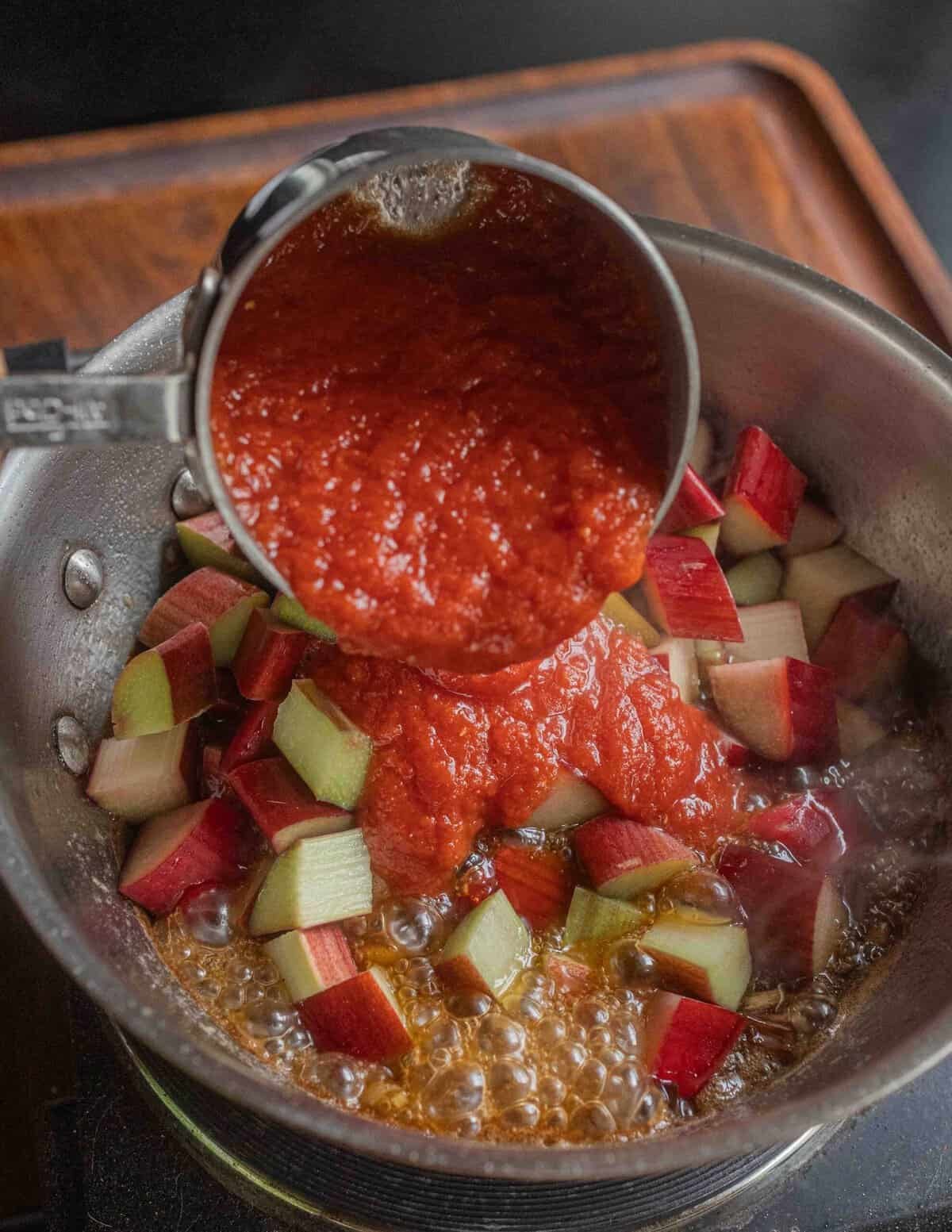 Adding tomato puree to a pan of rhubarb sauce on the stove.