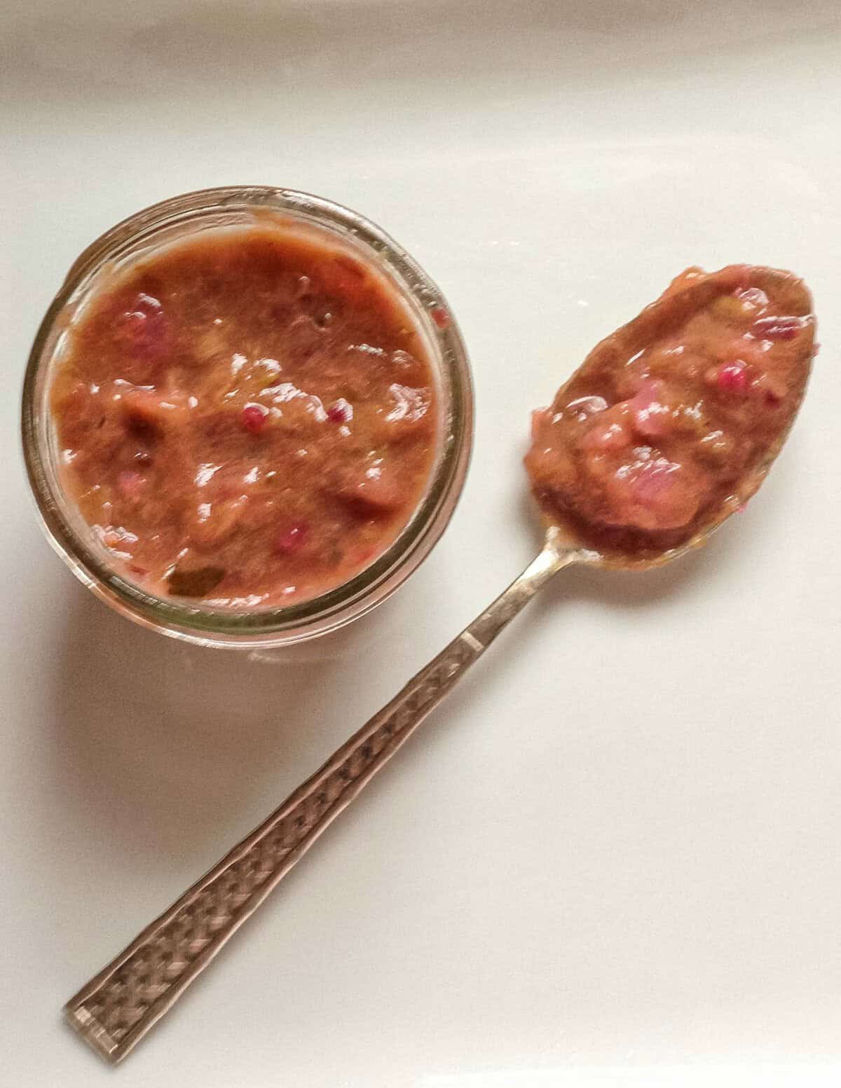 A small jar of rhubarb compote with a spoon on a white restaurant plate.