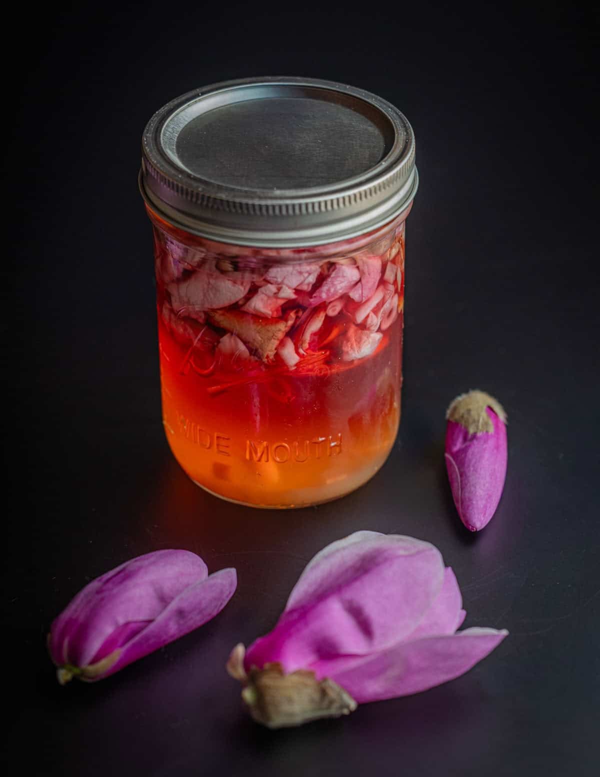 A jar of pickled magnolia petals surrounded by fresh magnolia flowers. 