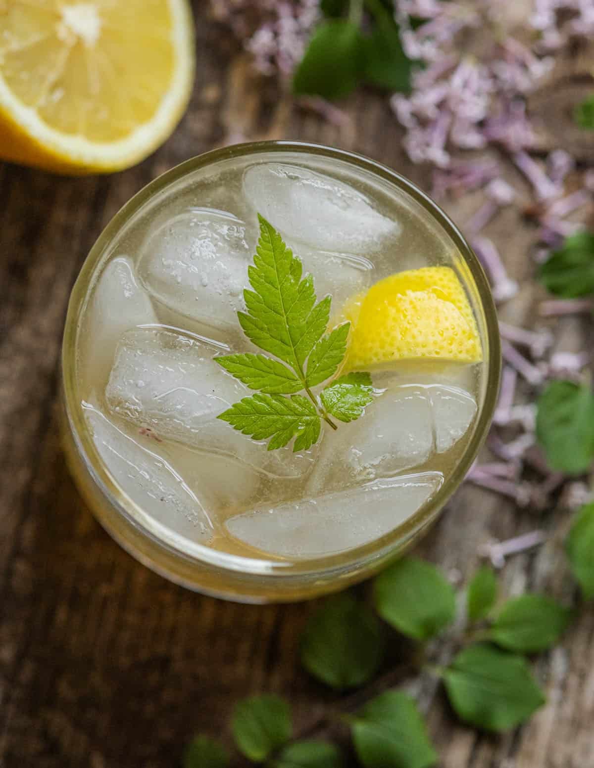 A glass of lilac lemonade seen from the top down showing lilacs around the glass and a leaf of anise root as a garnish.