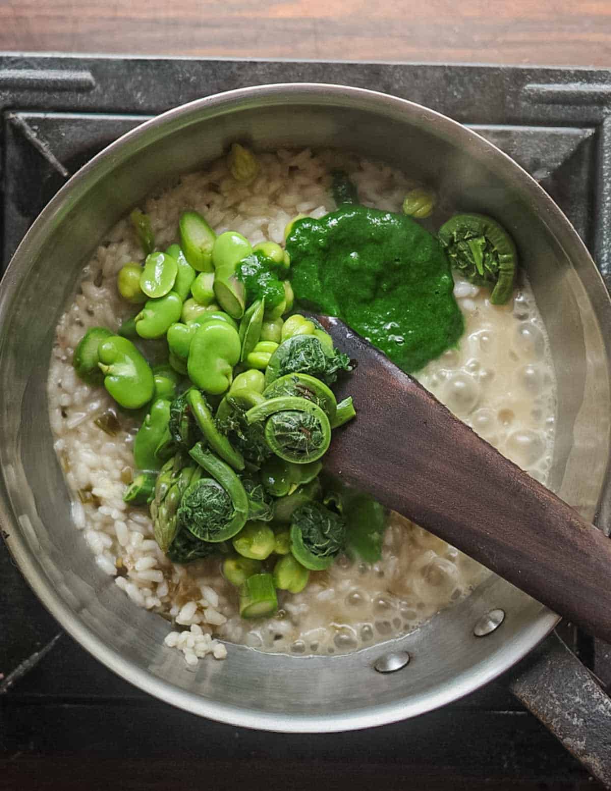 Adding blanched vegetables and ramp leaf puree to a pot of risotto.