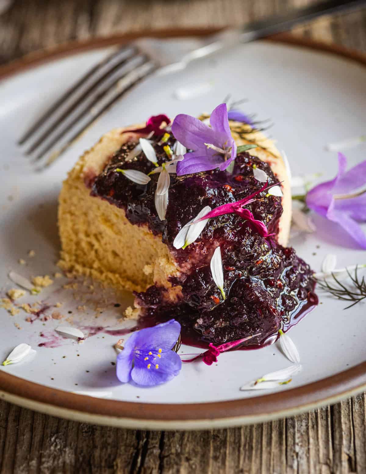 A cheesecake with fresh juneberry sauce being eaten garnished with edible bee balm, spiderwort, and campanula flowers.