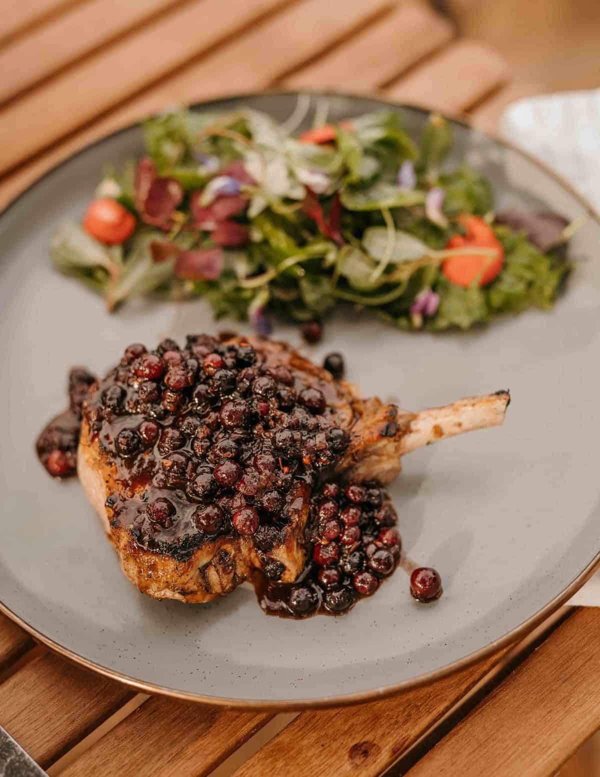 A pork chop on a plate topped with a ripe serviceberry or juneberry sauce with a wild green salad in the background.