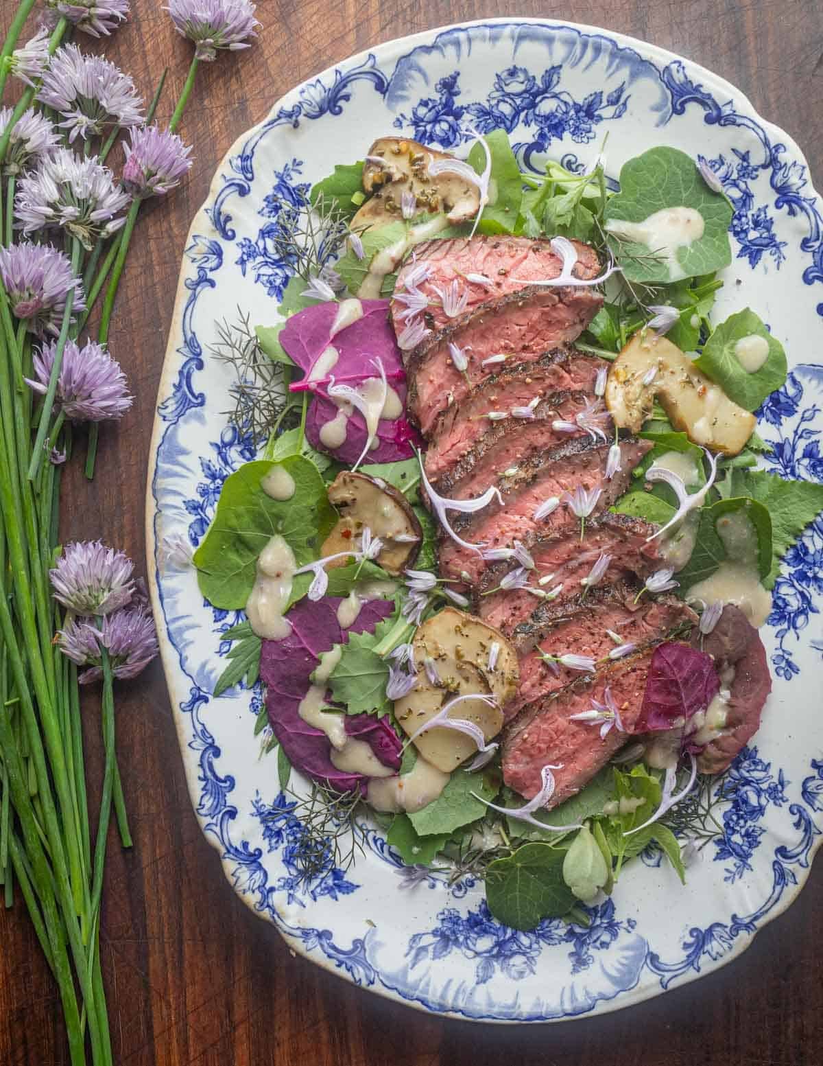 Sliced flank steak tagliata on a bed of fresh greens including orache and lambsquarters, drizzled with chive blossoms vinaigrette and garnished with fresh chive blossoms.