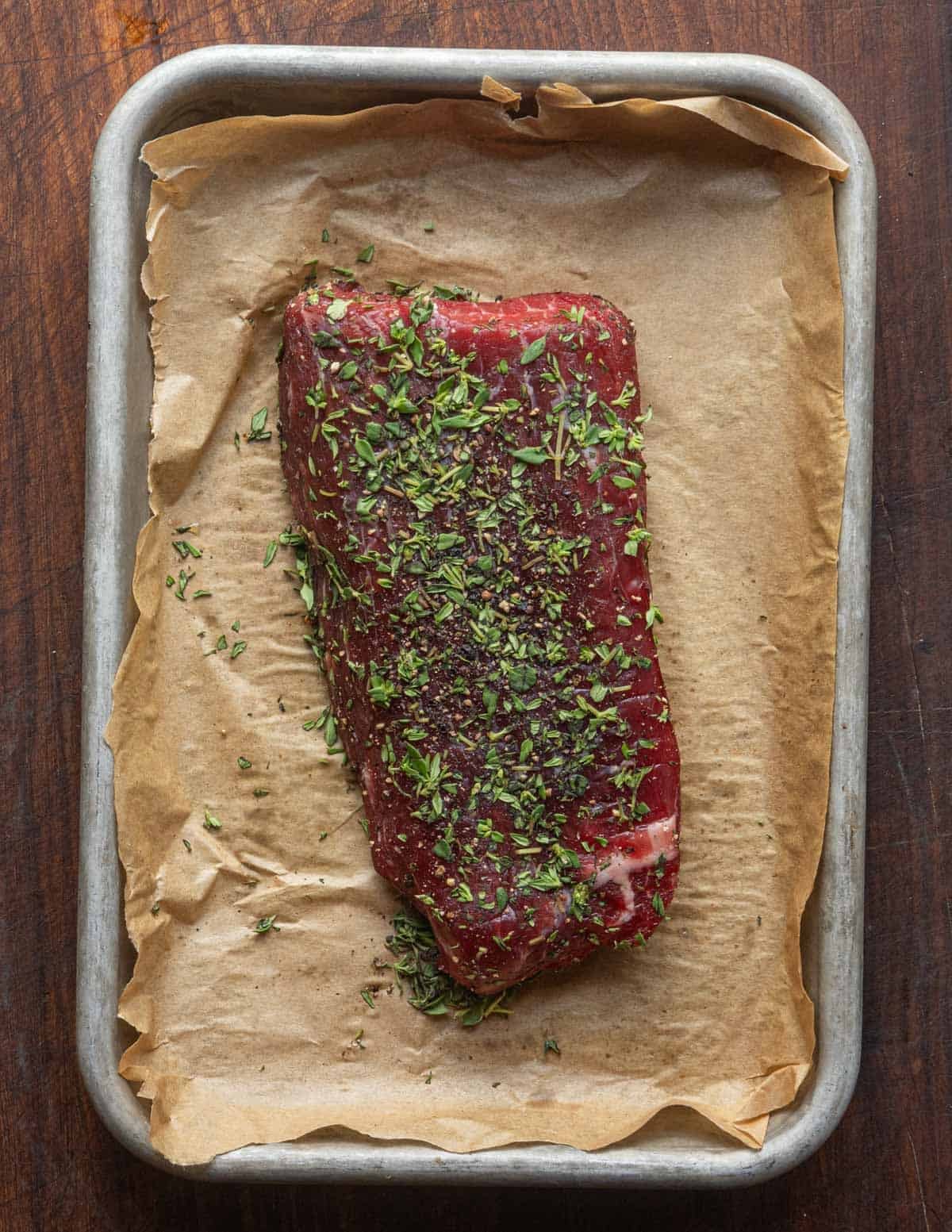 A flank steak seasoned with salt, pepper and thyme, on a baking sheet lined with parchment.