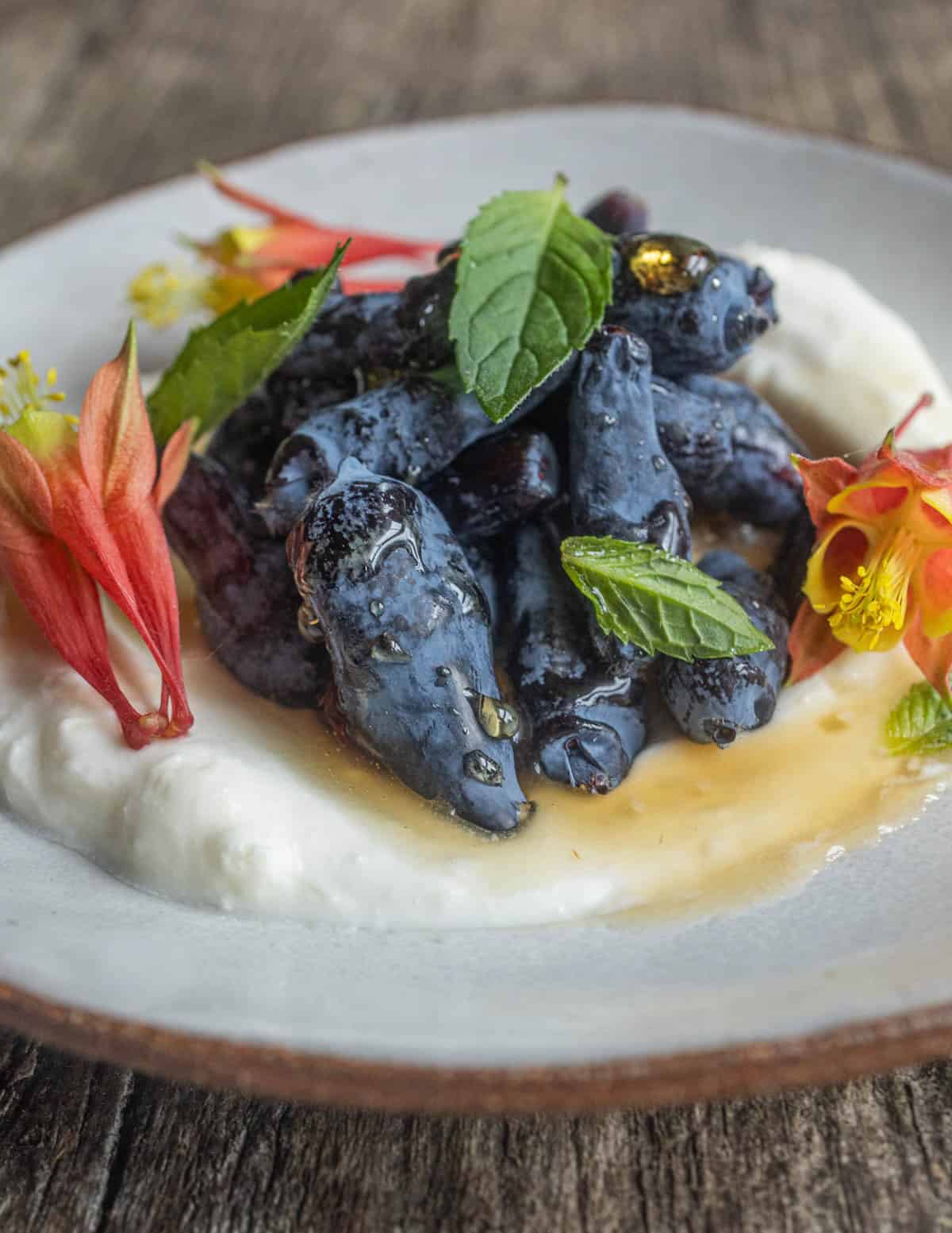 A small plate with fresh honeyberries on a bed of creme fraiche, with maple syrup, mint leaves and columbine flowers.