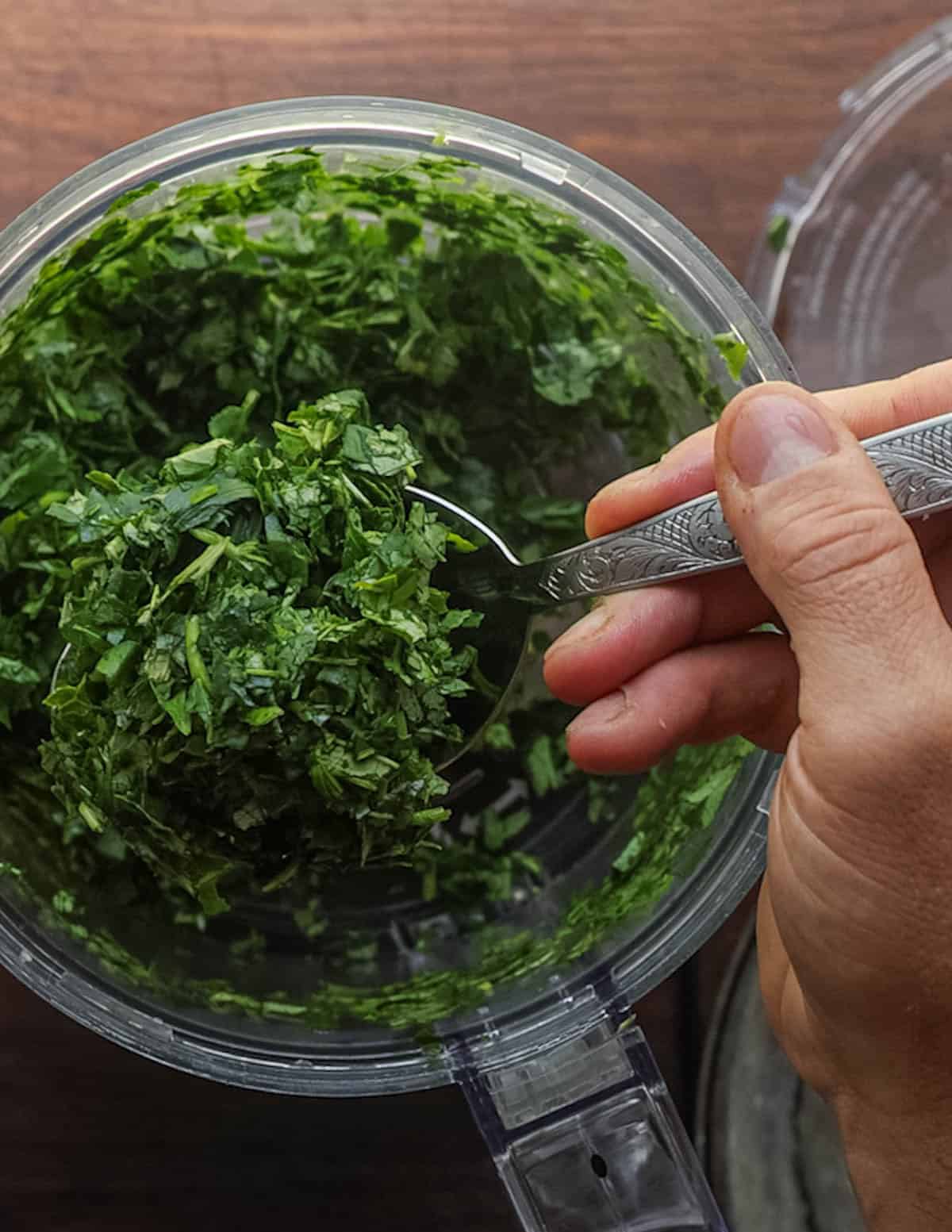 A hand holding a spoon filled with finely chopped sweet potato leaves from a food processor.