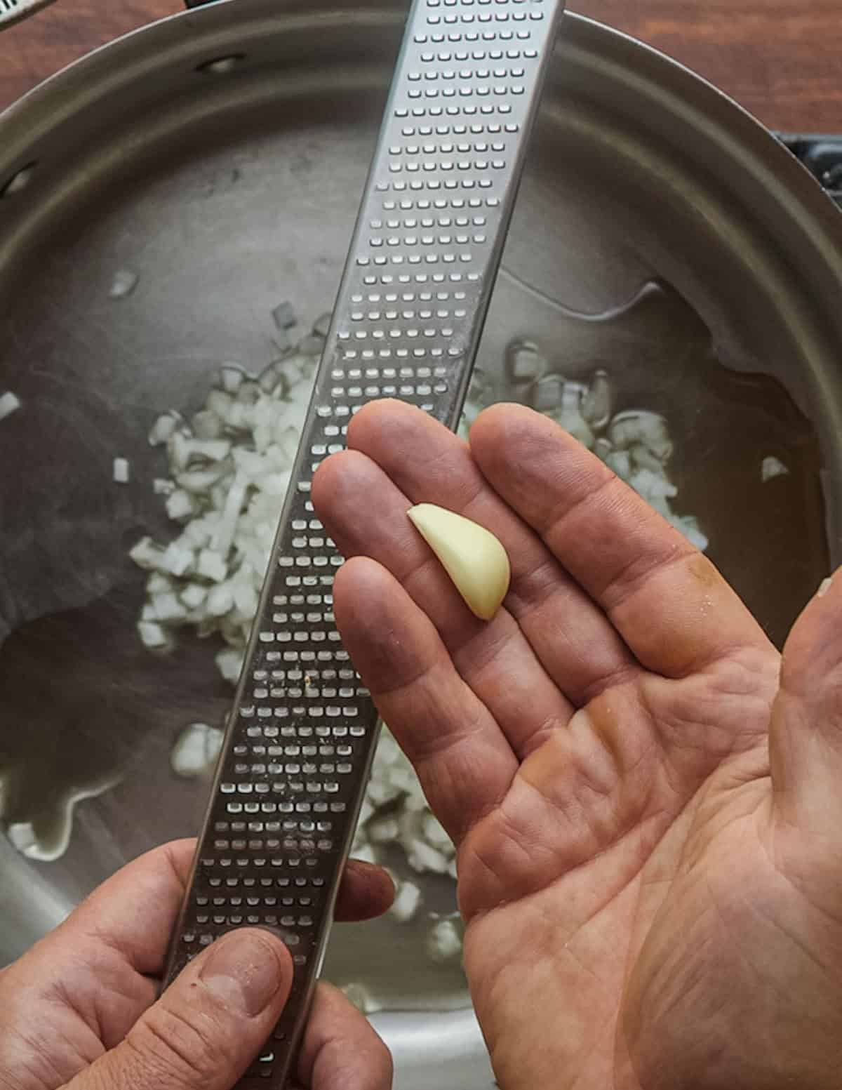 A hand seen over a pan with cooking onions grating a garlic clove on a microplane grater.