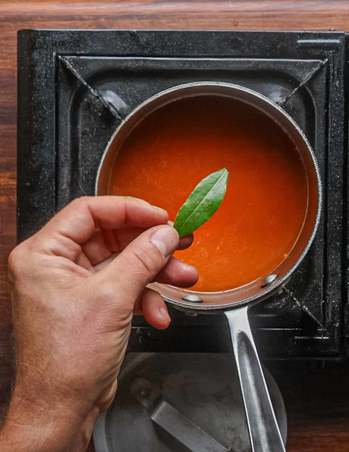A hand laying a fresh bay leaf into a pot of rice cooking in tomato paste and water.
