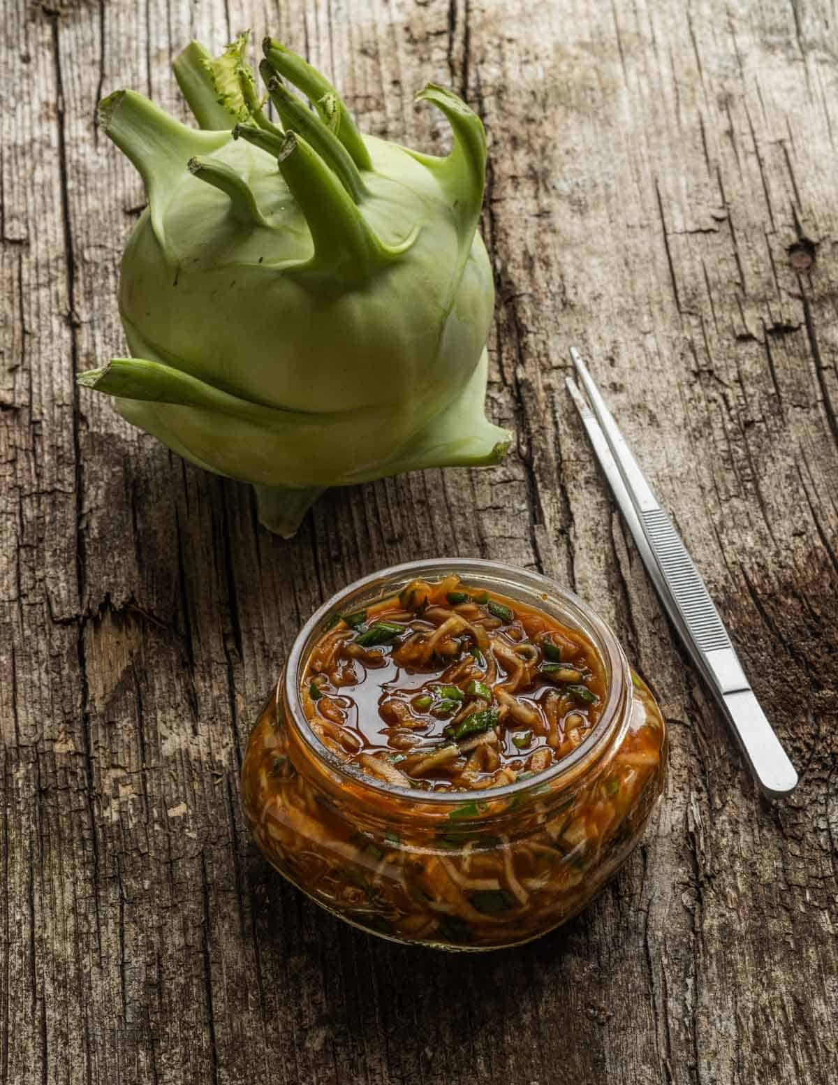 A jar of fermented kohlrabi kimchi next to a whole kohlrabi bulb and a tweezers for removing the kohlrabi.