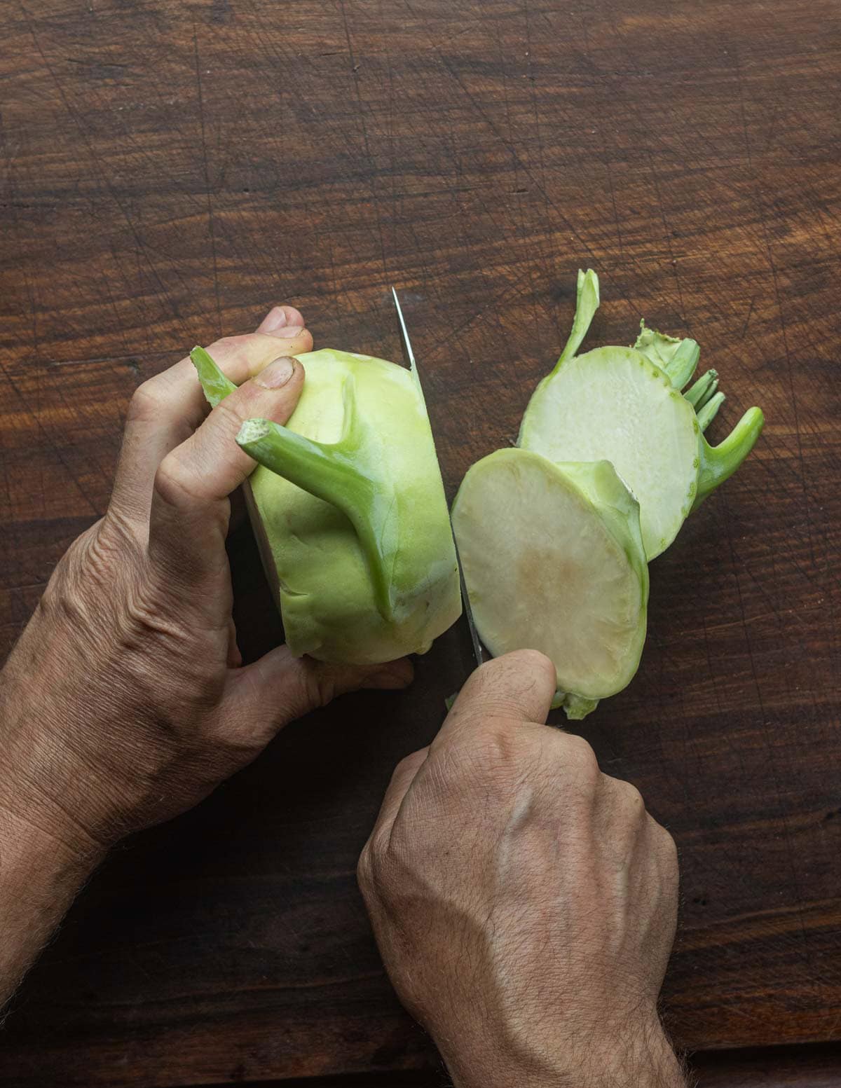 A point of view image looking down on two hands cutting the top and bottom of a kohlrabi bulb off.