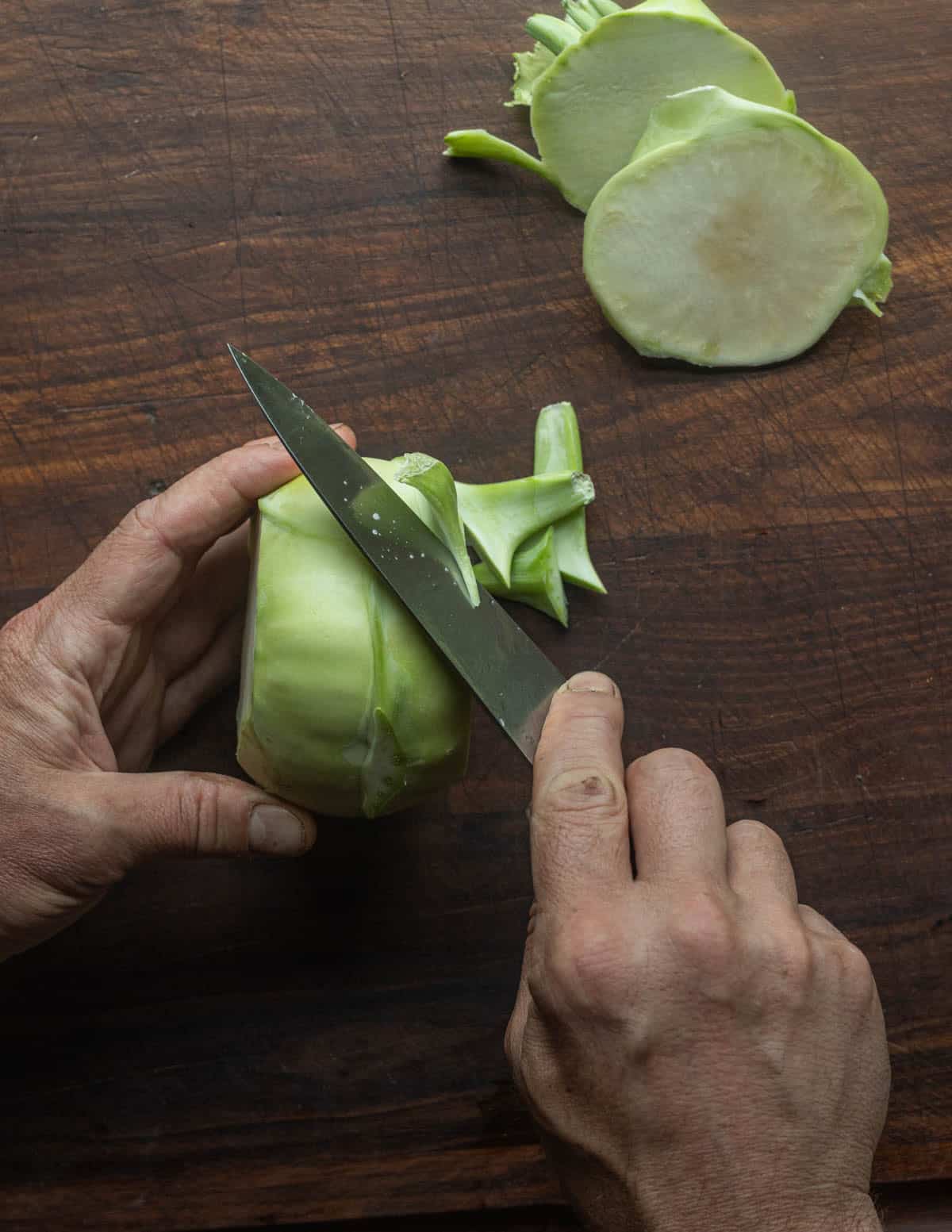 A top down image showing two hands cutting off the remaining stem pieces of a kohlrabi bulb.