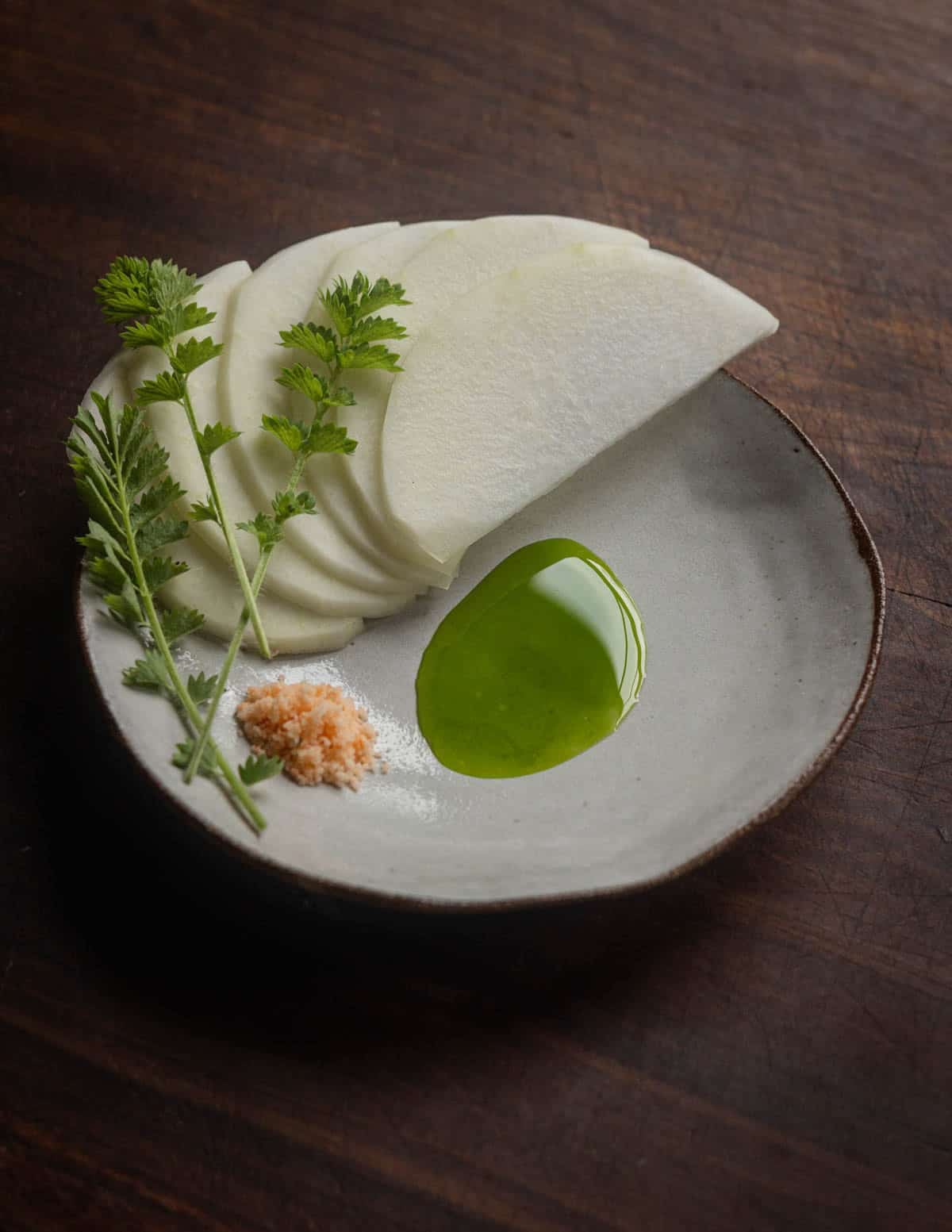 Sliced raw kohlrabi on a plate with herb oil, ghost pepper salt and sprigs of burnet.