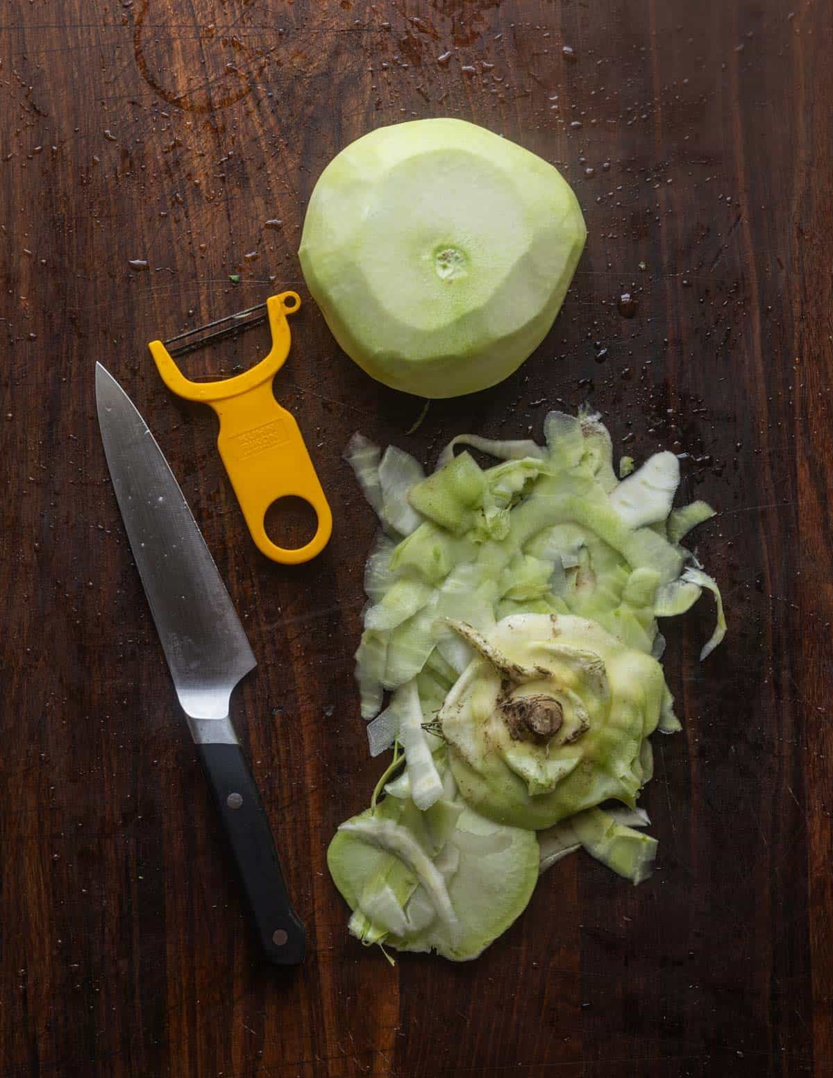 A finished, peeled bulb of kohlrabi on a cutting board next to a y-shaped peeler and a chef's knife.