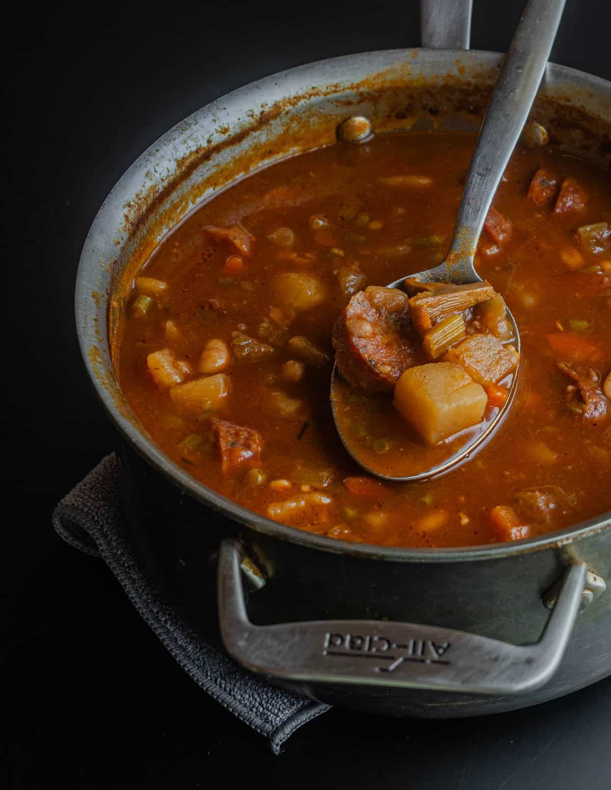 A pan of sausage, vegetable and kohlrabi soup in an all clad pot on a trivet.