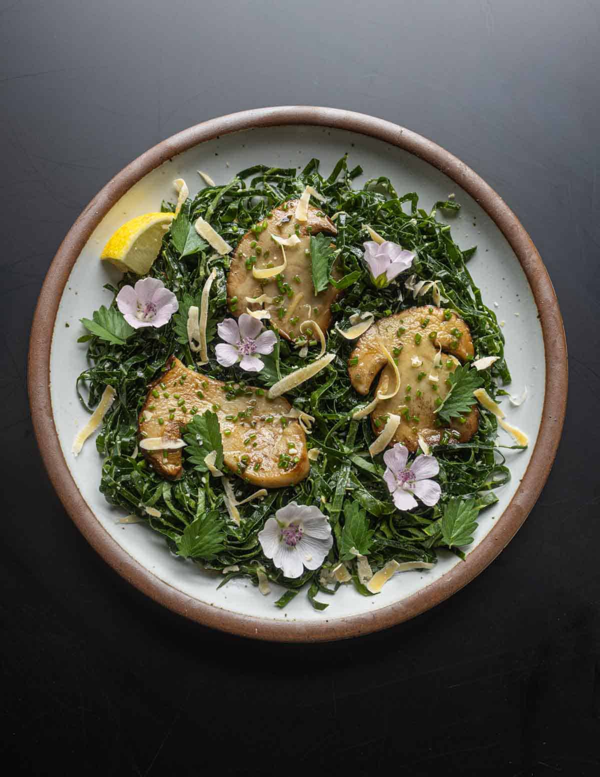 A top down image of a shredded kohlrabi greens salad seasoned with Caesar vinaigrette and garnished with sliced boletus rex veris mushrooms, burnet leaves and wild pine nuts.