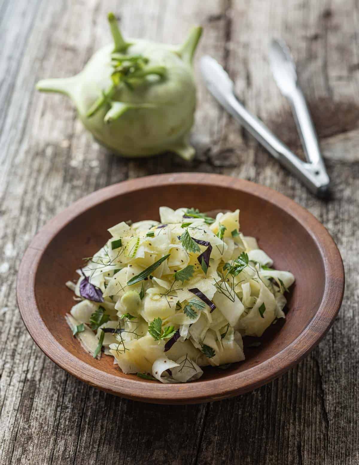 Shaved kohlrabi, apple and fennel salad with burnet, tarragon, chives and opal basil in a wooden bowl with a kohlrabi bulb in the background and a set of serving tongs.