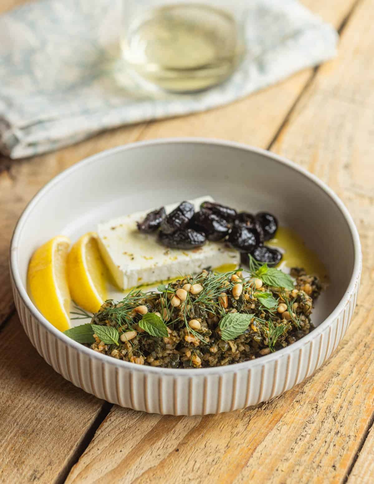A round plate with a tall rim with a serving of Greek spinach rice with feta cheese, black olives, olive oil and lemons with a glass of white wine in the background.