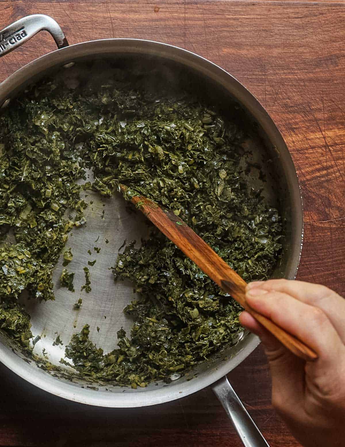 A hand drawing a spatula through a pan of cooked wild greens showing the bottom of the pan which indicates it's properly cooked.