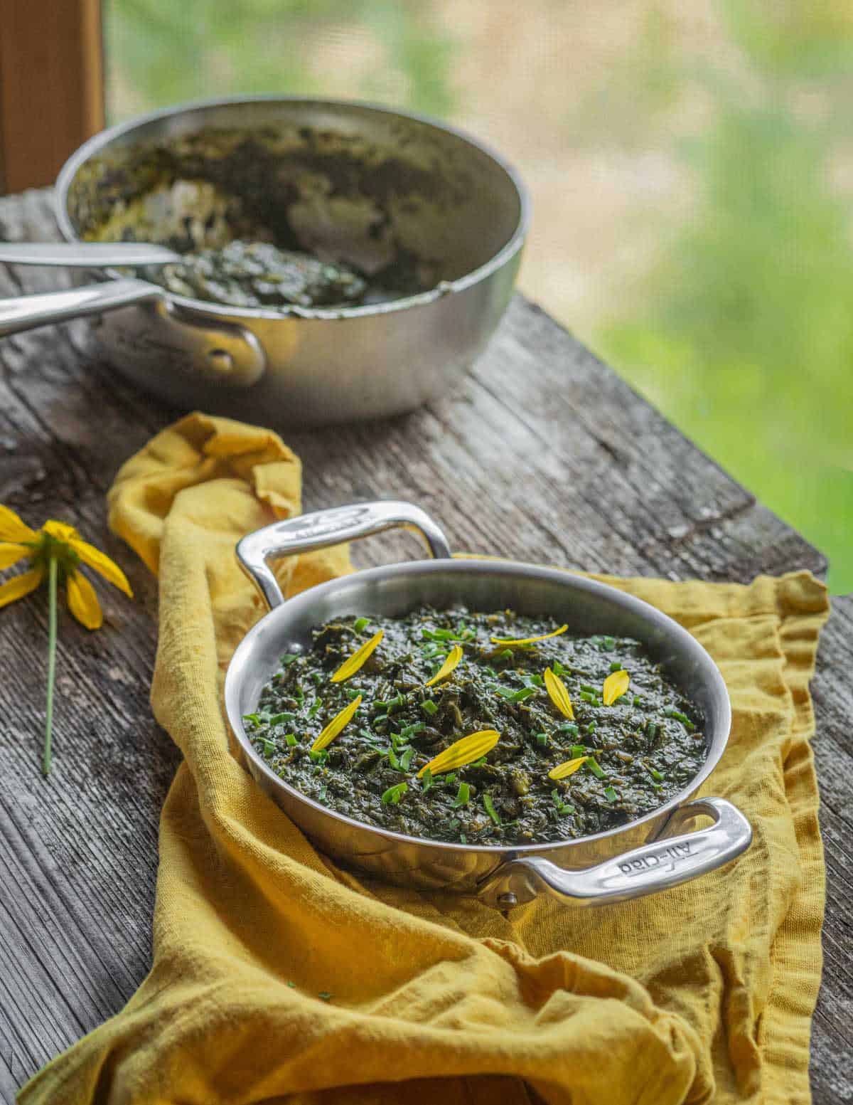 A small All Clad pan filled with a serving of African sweet potato vine stew on a yellow napkin with a cooking pan in the background.