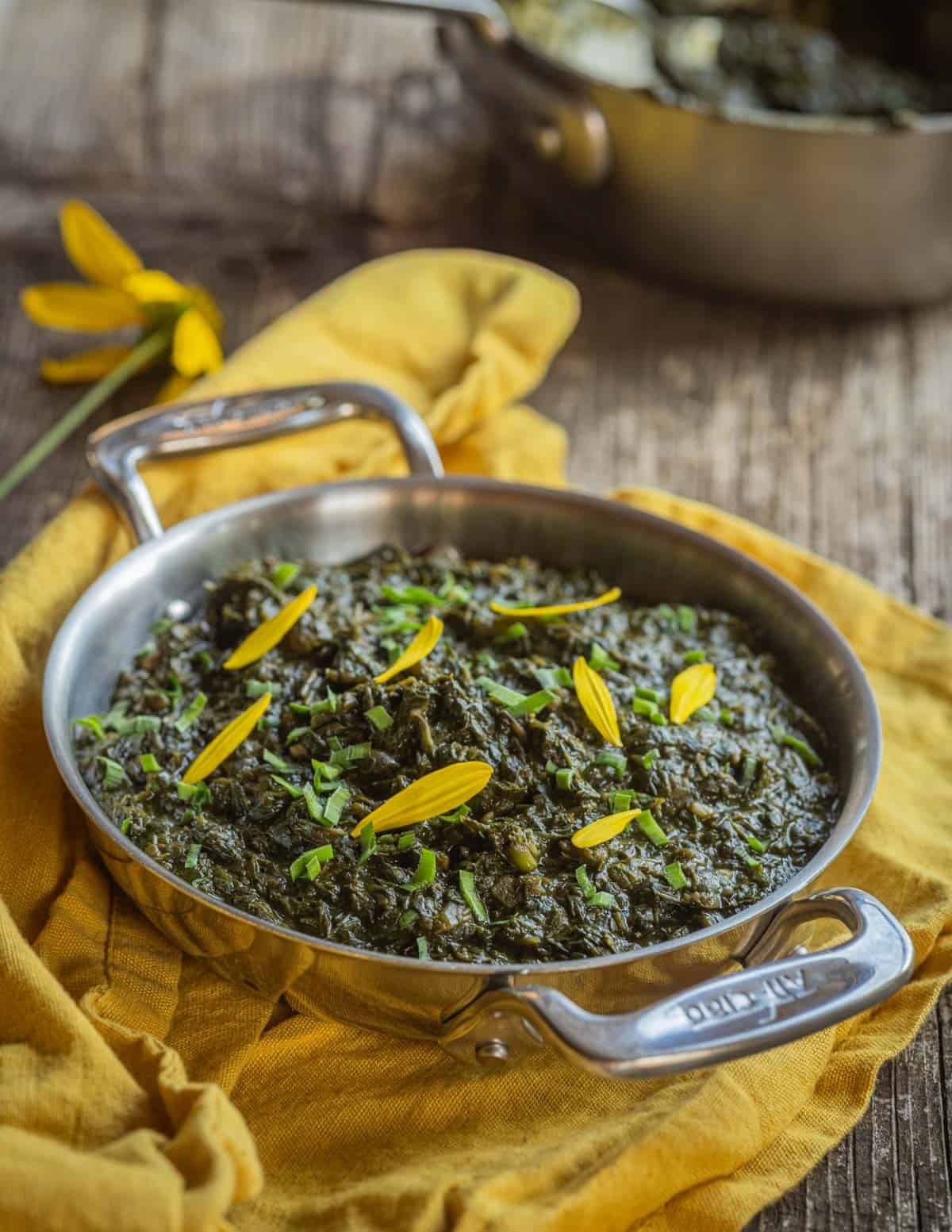 A small All Clad dish of Guinean sweet potato leaf stew garnished with yellow rudbeckia flowers and a yellow napkin.