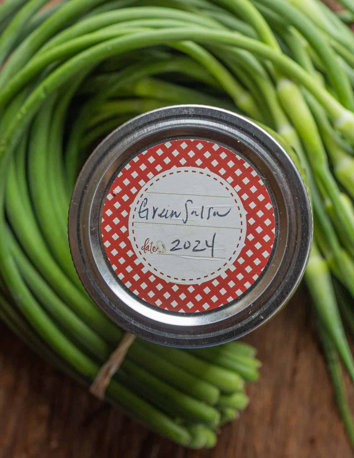 A jar of homemade garlic scape salsa surrounded by fresh garlic scapes.