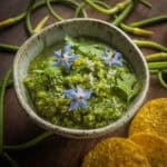 A bowl of tomatillo and garlic scape salsa garnished with borage flowers in a bowl surrounded by fresh garlic scapes and tortilla chips.