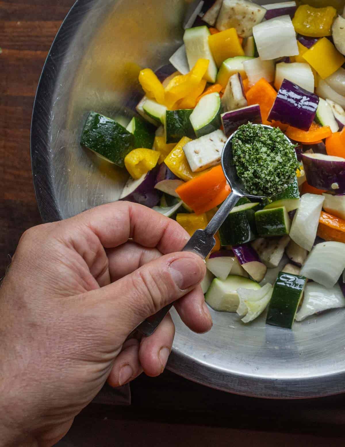 A bowl of diced eggplant, onion, peppers, and zucchini being seasoned with fresh herb salt.