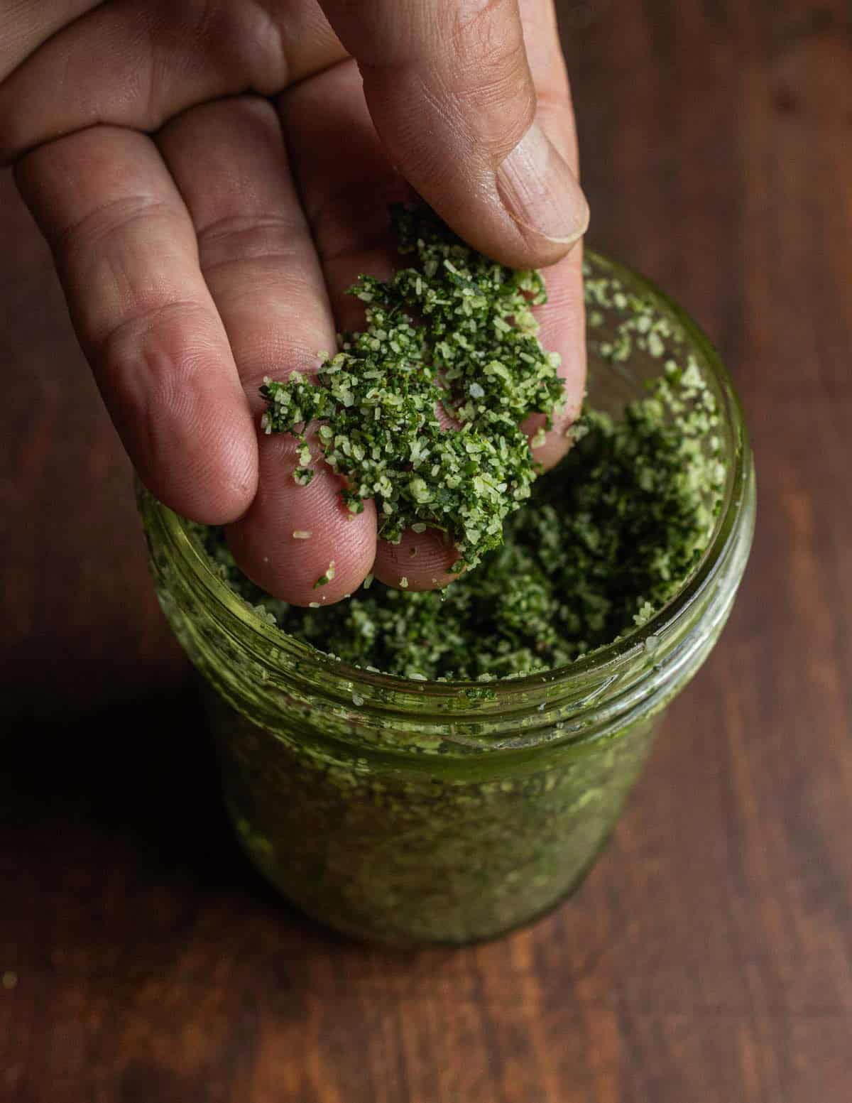 A hand holding a pinch of green herb salt from a mason jar showing the texture is similar to wet sand.
