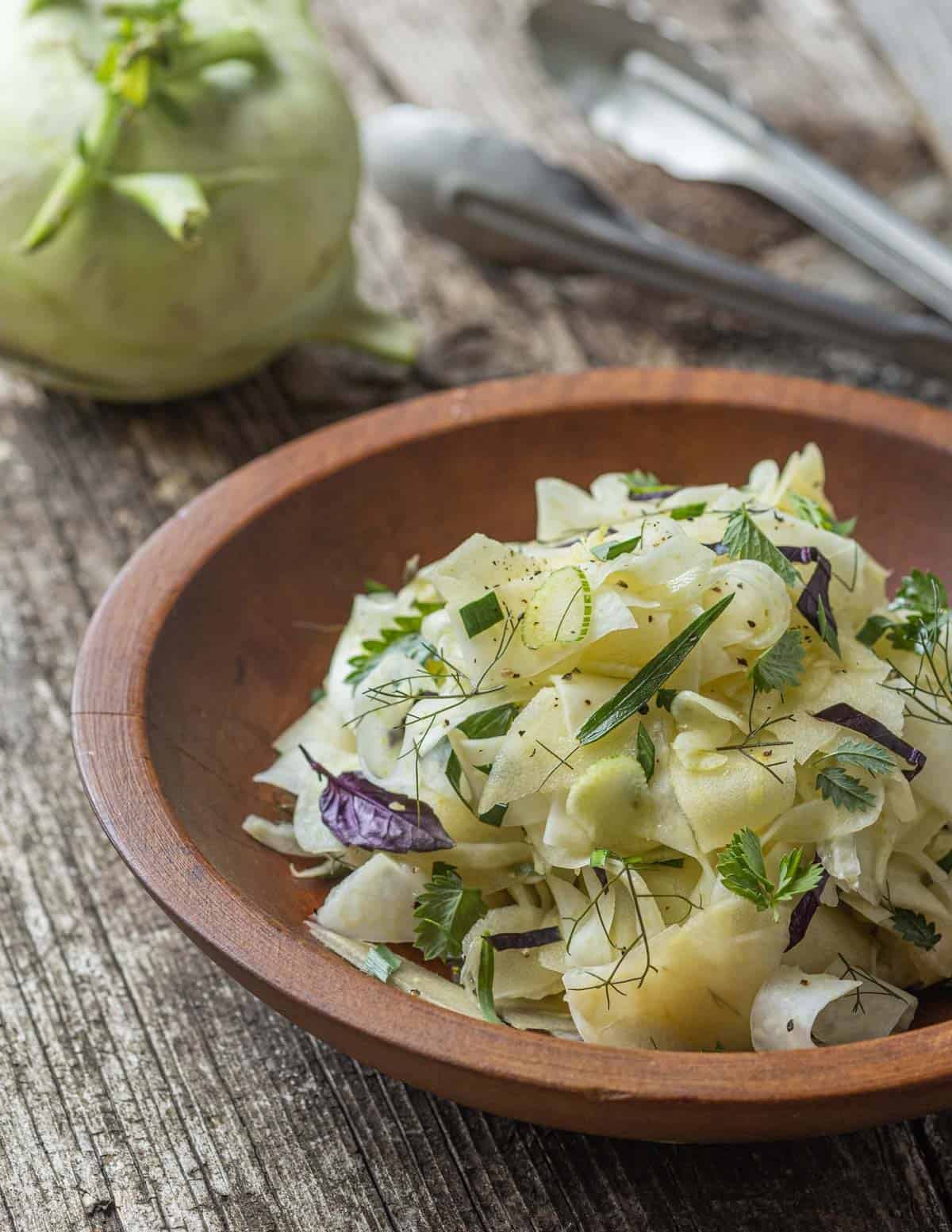 Kohlrabi, apple and shaved fennel salad with herbs in a wooden bowl with a kohlrabi bulb in the background.