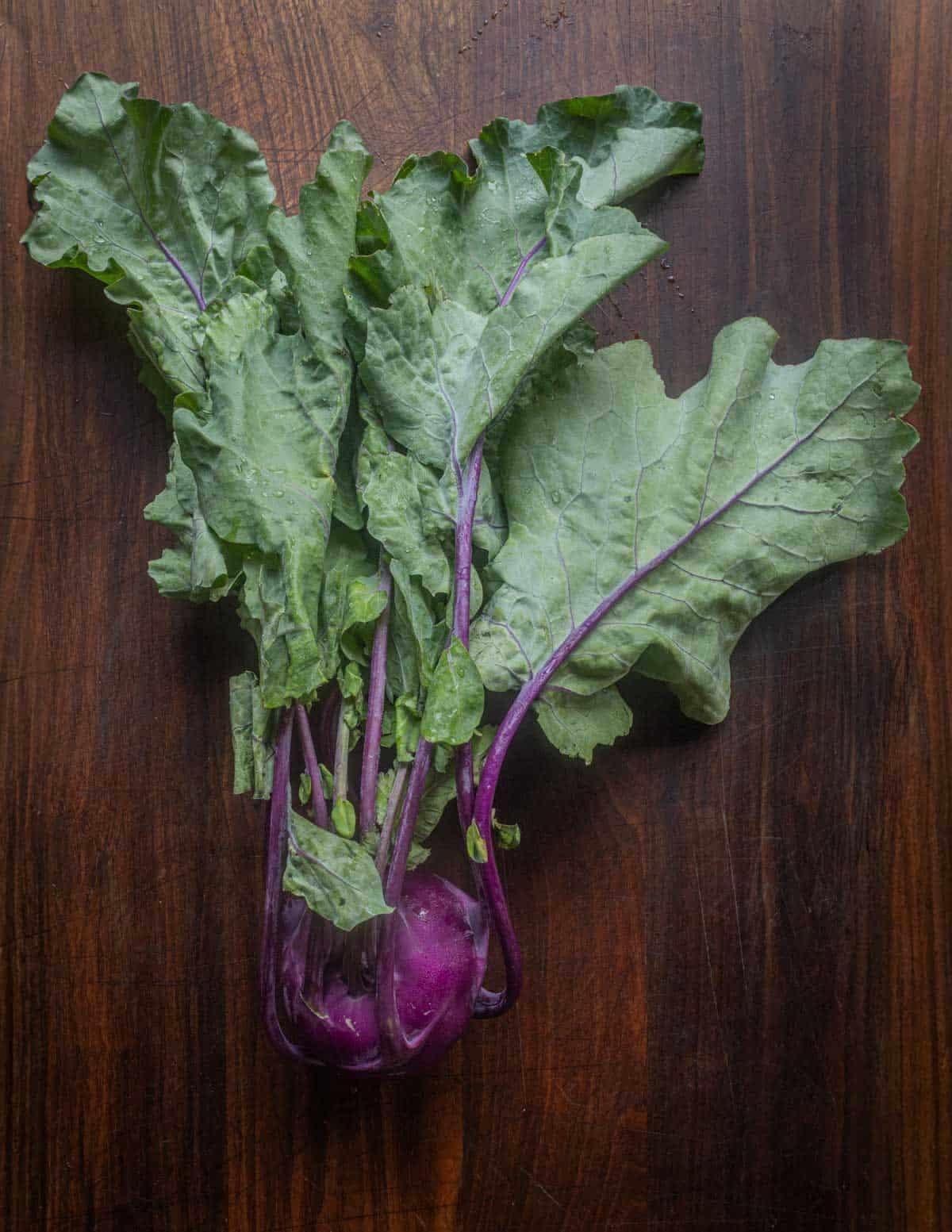 A purple kohlrabi bulb with attached greens on a walnut cutting board.