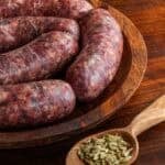 Hot Italian fennel sausage on a wooden plate next to a wooden spoon filled with whole fennel seeds.