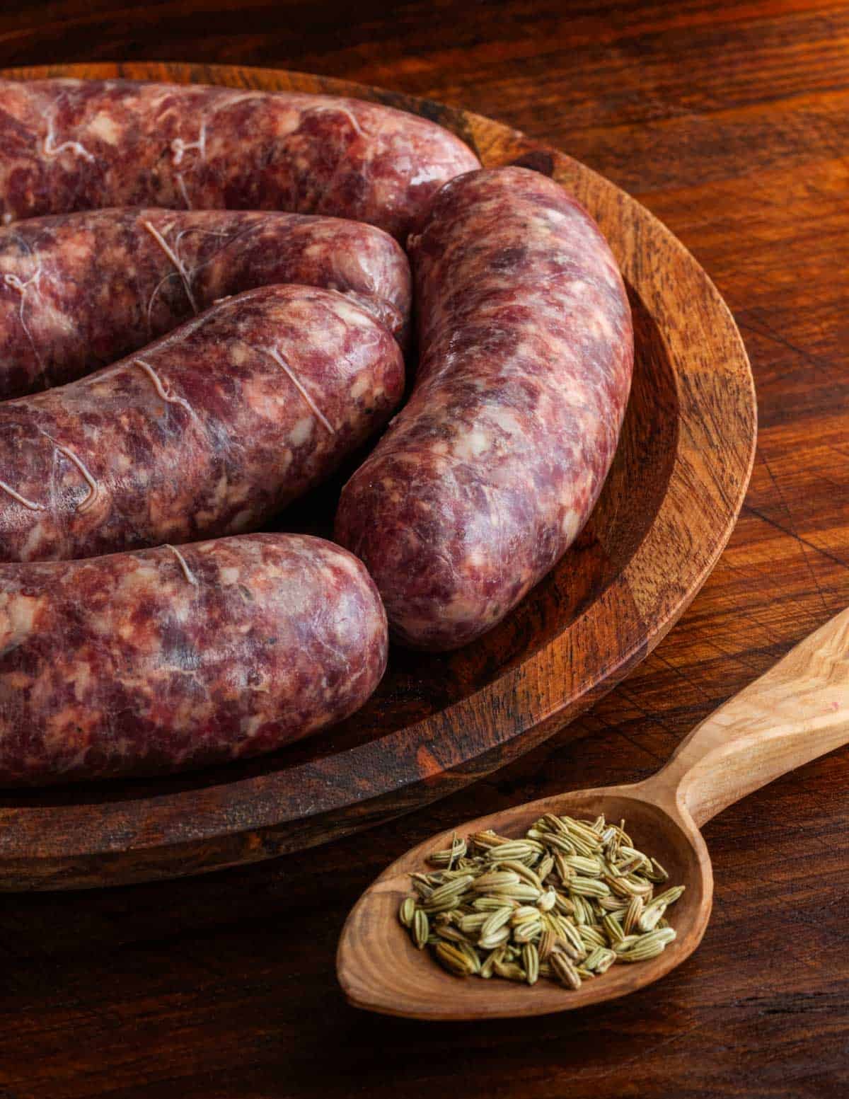 Hot Italian fennel sausage on a wooden plate next to a wooden spoon filled with whole fennel seeds.