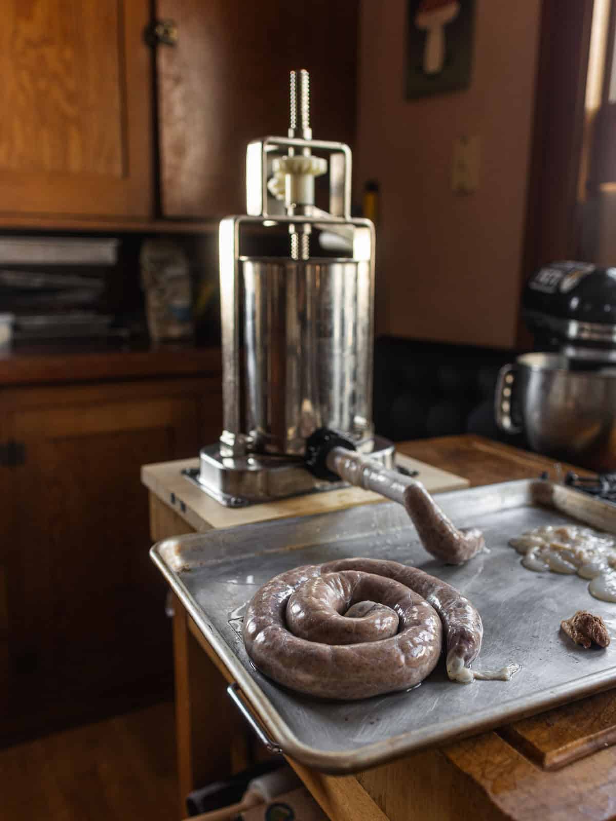 A sausage stuffer on a cutting board with filled sausages next to it on a sheet tray in a home kitchen.