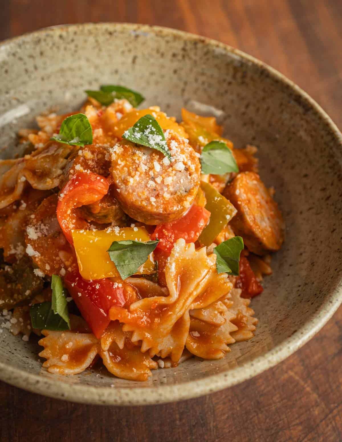 A bowl of farfalle pasta with sausage, peppers, tomato and basil garnished with pecorino or parmesan cheese.