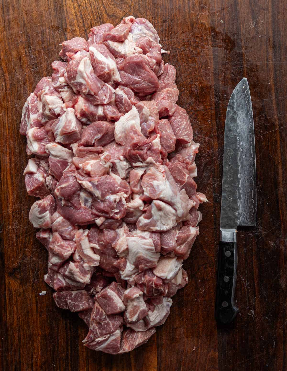 A pile of diced pork shoulder on a cutting board next to a chef's knife.