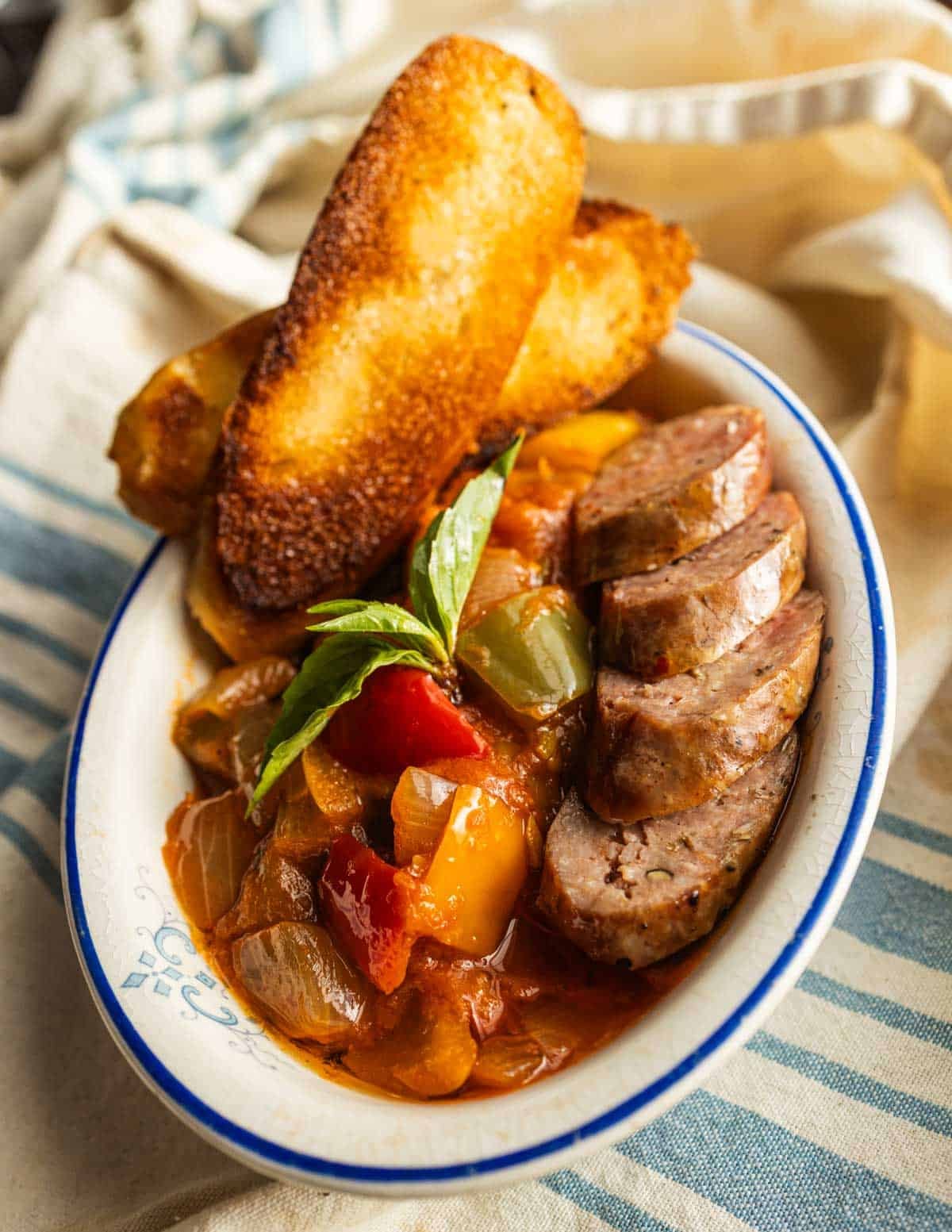 A bowl of peperonata with grilled, sliced Italian sausage, toasted bread and basil leaves.