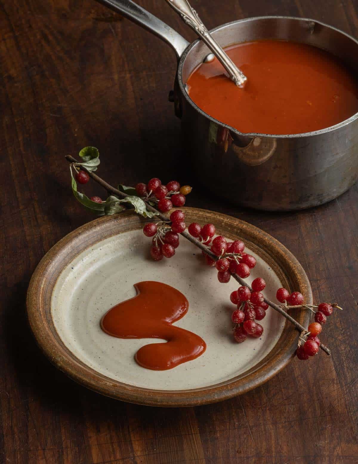 Autumn olive ketchup on a plate next to a branch with ripe fruit and a pan of red sauce in the background.