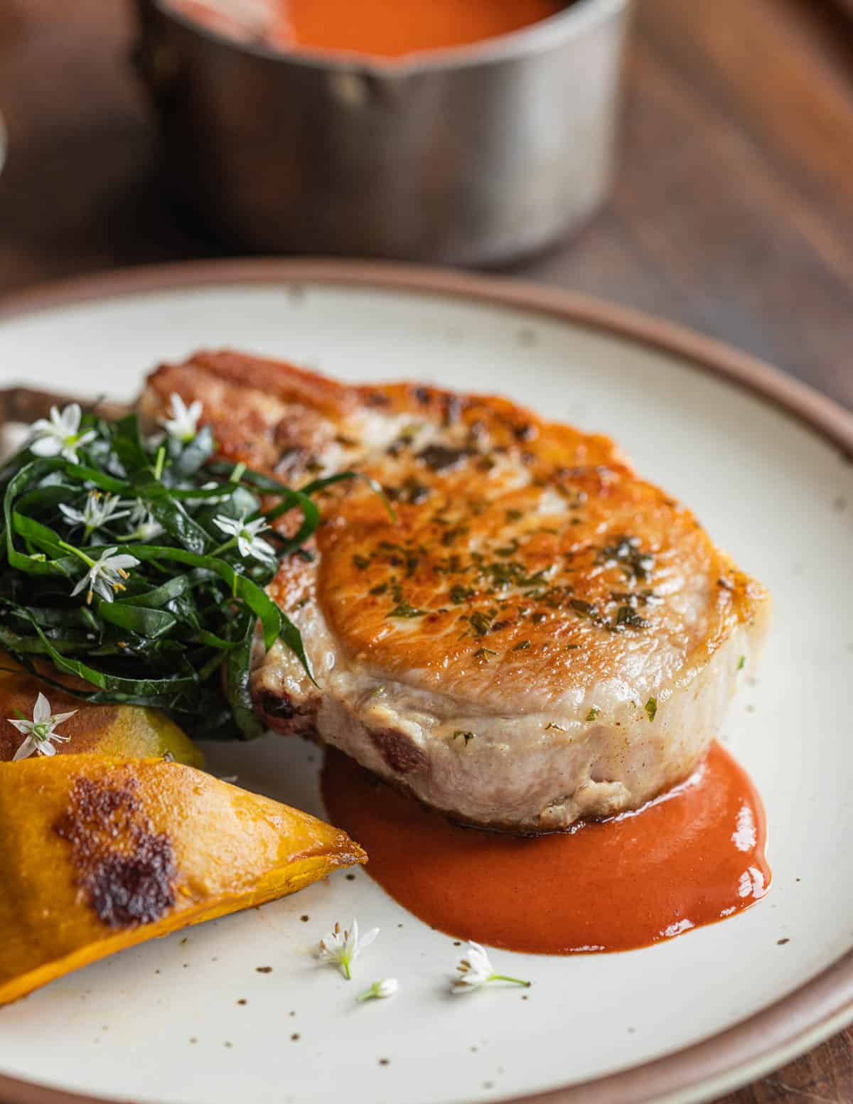 A bone in pork chop on a plate with squash, broccoli greens, garlic chive flowers and autumn olive ketchup sauce.