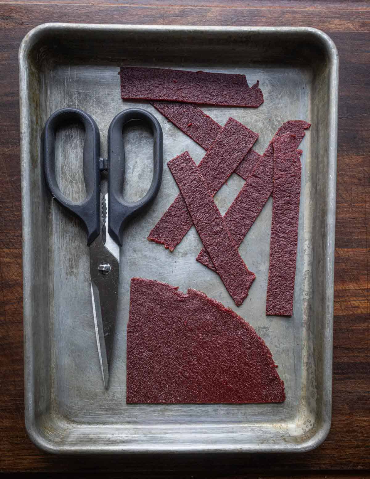 A baking sheet with a kitchen shears being used to cut autumn olive fruit leather. into strips.